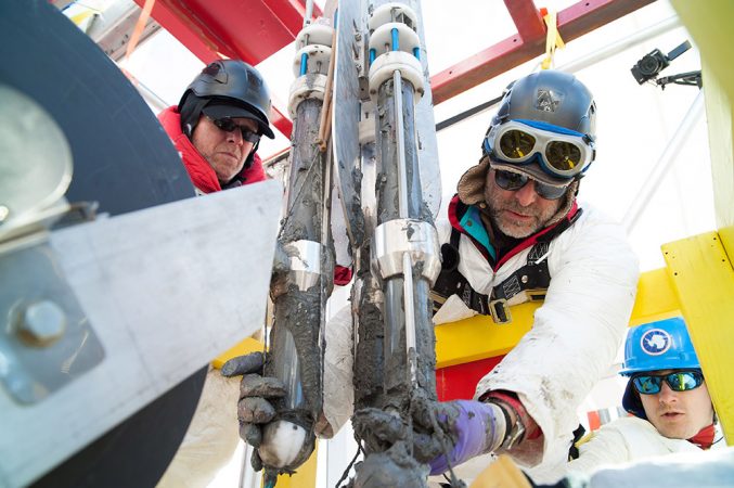 As Ross Powell (on left) looks on, Reed Scherer (muddy hands) takes hold of the corer. It has just emerged from Lake Whillans full of mud. Scherer and Powell wear climbing harnesses that are connected to the metal frame above — just in case they slip and fall into the half-mile-deep hole beneath their boots! Credit: © JT Thomas