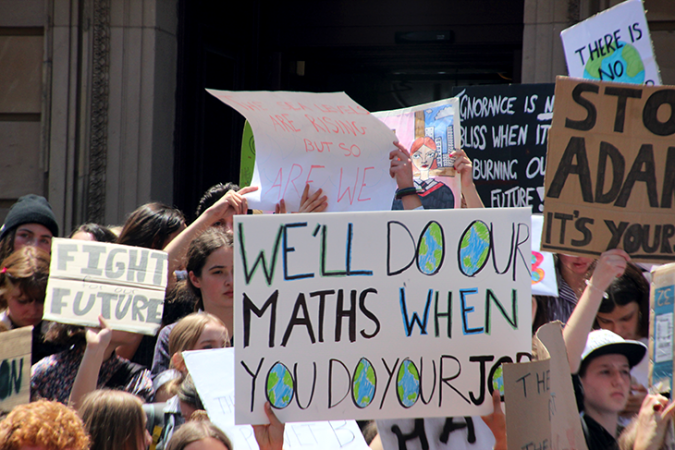 a photo of a student climate strike in Australia, a sign in the center of the picture reads "We'll do our maths when you do your job". All of the Os in the message are planet Earths.