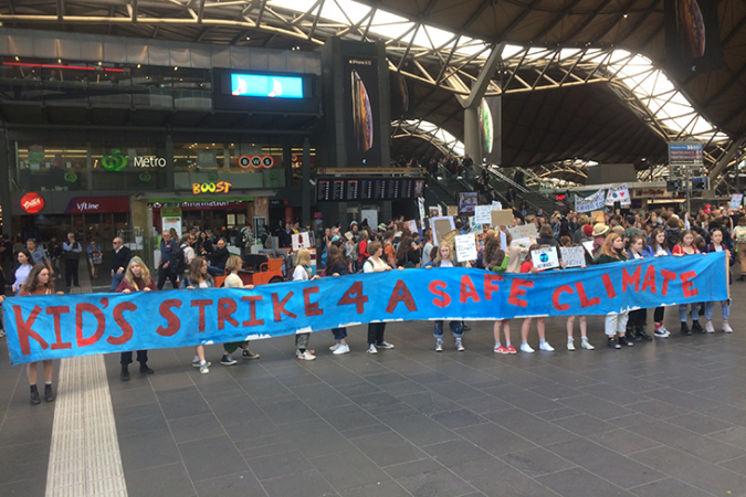 a photo of a climate strike in Australia, a row of students holds a long blue banner with red lettering that reads "Kid's Strike 4 A Safe Climate"