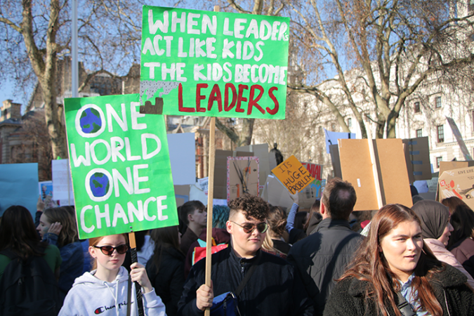 A photo of a student strike in London showing two green banners. One reads "One World Once Chance", and the other reads "When Leaders Act Like Kids The Kids Become Leaders"