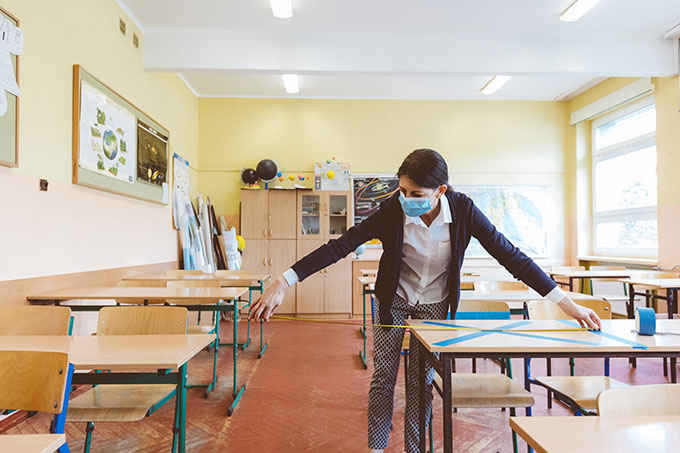 a photo of a masked teacher measuring distance between desks with her arms