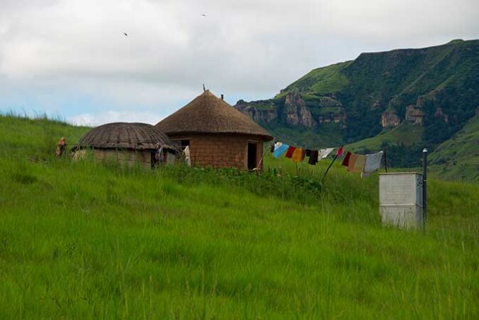 a photo of a grey outhouse on a grassy hill next to a family's home