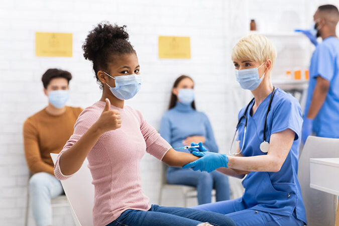 a masked teen getting a shot looks at the camera and gives a thumbs up