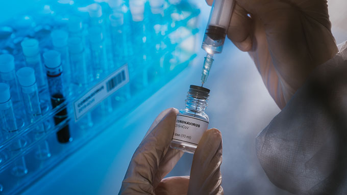 gloved hands hold a syringe with needle and a vial with the label COVID-19 vaccine