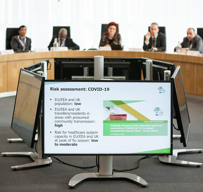 a photo of people sitting at a curved table behind monitors showing COVID-19 prediction data