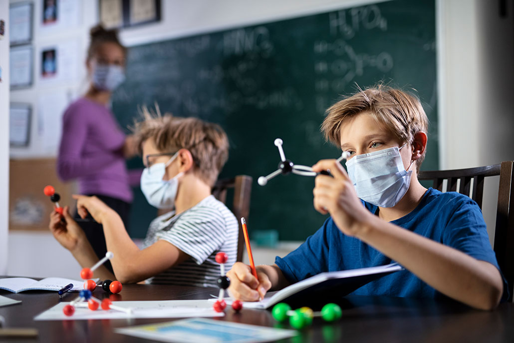 two boys look at molecular models and a teacher writes on a chalkboard behind them