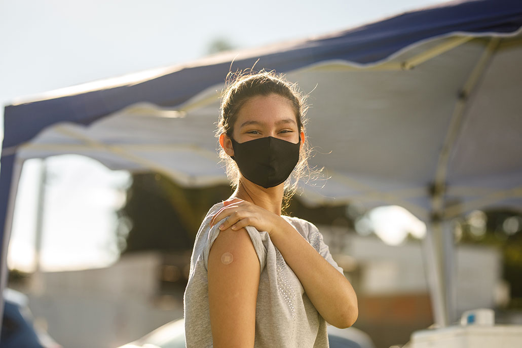 a girl standing outside showing a bandaid on her upper arm and wearing a face mask