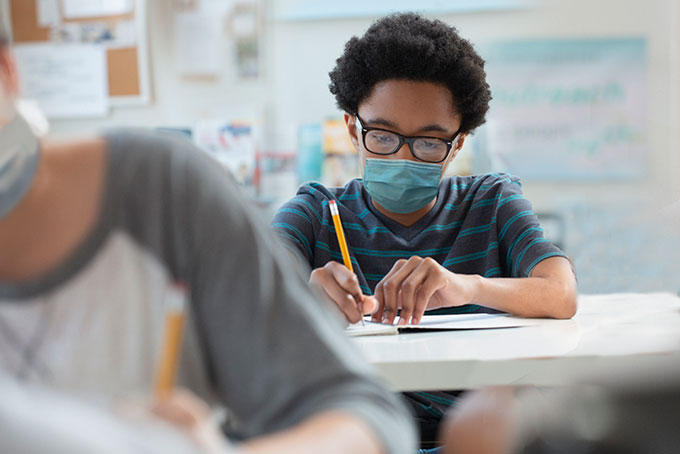 a kid sitting at a school desk wearing a mask