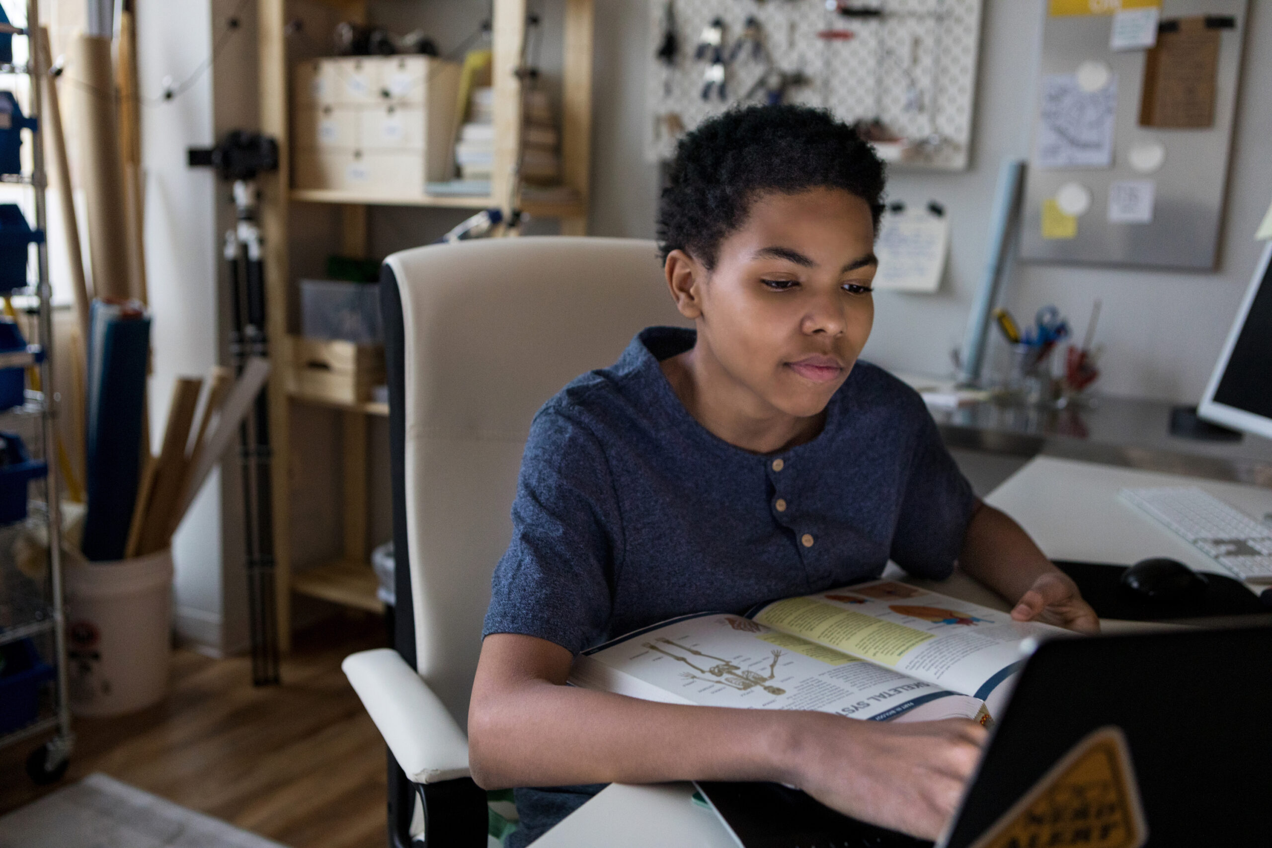 a boy sits at a bedroom desk with a book and laptop open in front of him