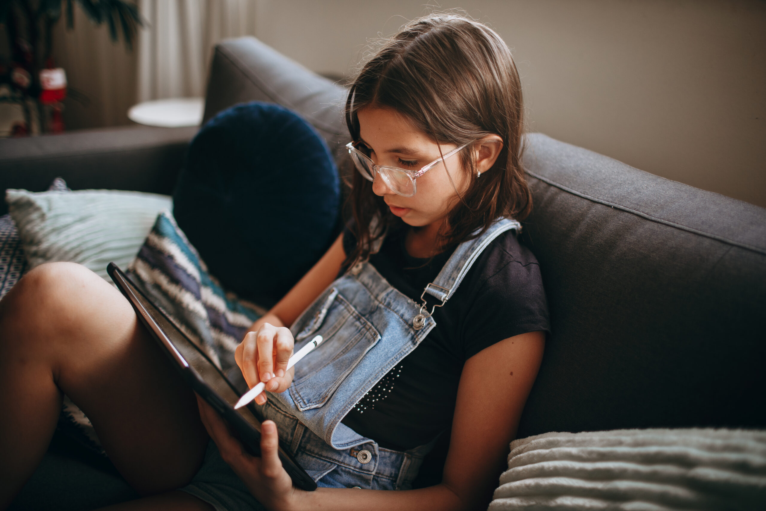 a girl wearing overalls and glasses sits on a couch and reads on a tablet screen