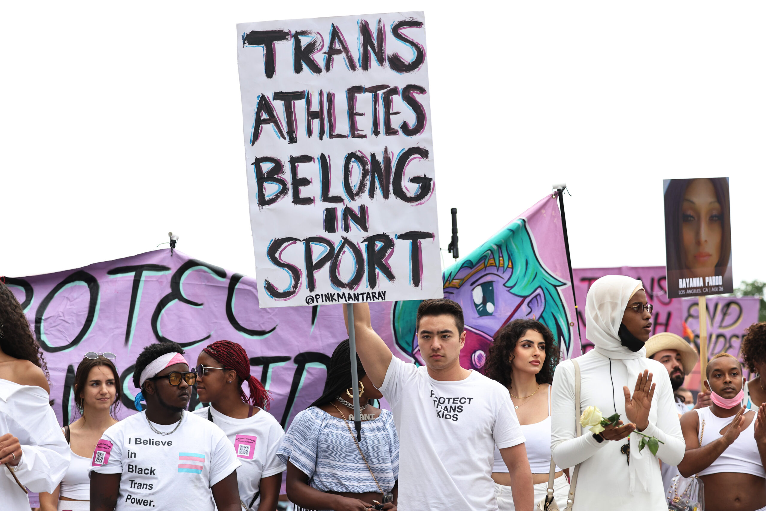 activists protest for transgender rights, one holding a sign that reads 'trans athletes belong in sport'