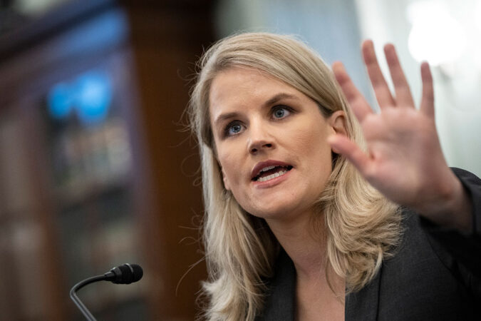 a photo of Frances Haugen, a white lady with blond hair, holding her hand up during her testimony