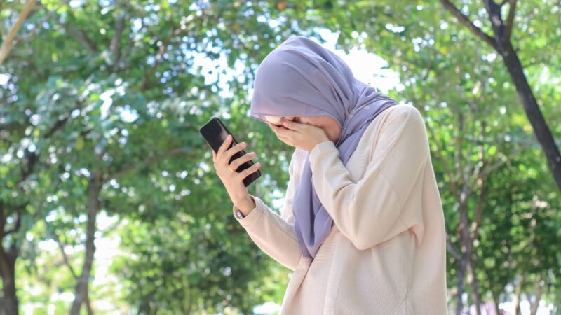 a Muslim woman wearing a headscarf holding a phone and covering her face, She looks sad.