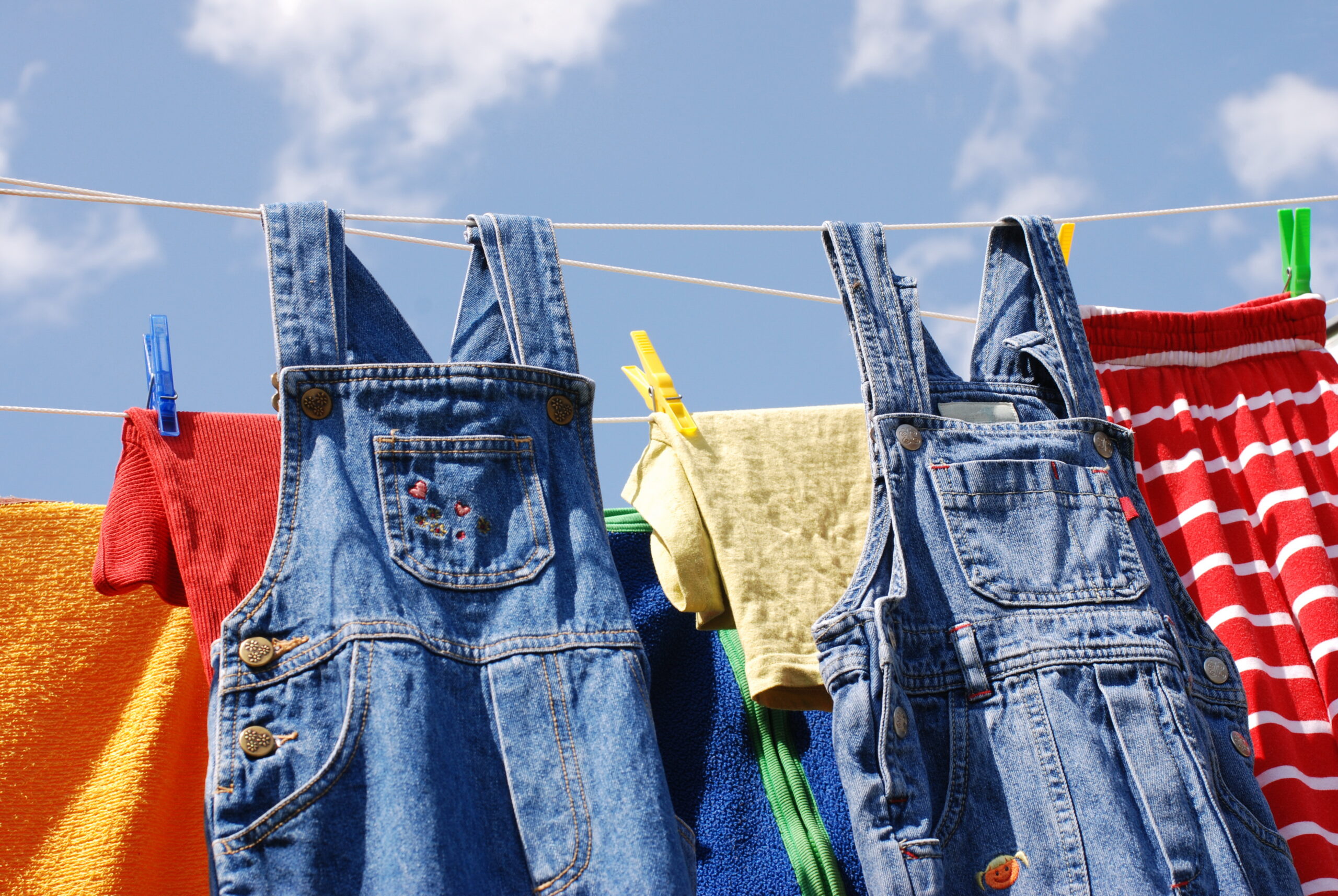 two pair of denim overalls hang out on a clothes line under a blue sky with other garments