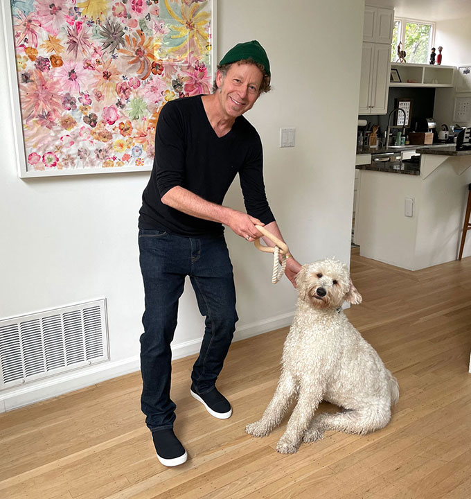 Ken Goldberg stands inside next to his dog Rosie. Rosie is a medium to large sized dog with curly white fur. Ken is holding a pull toy.