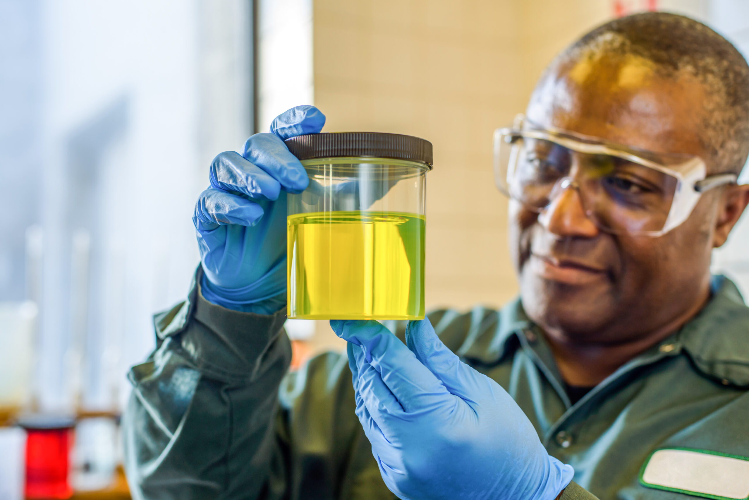 an adult Black man holds a clear canister containing a yellow liquid, while wearing blue rubber gloves and protective goggles 