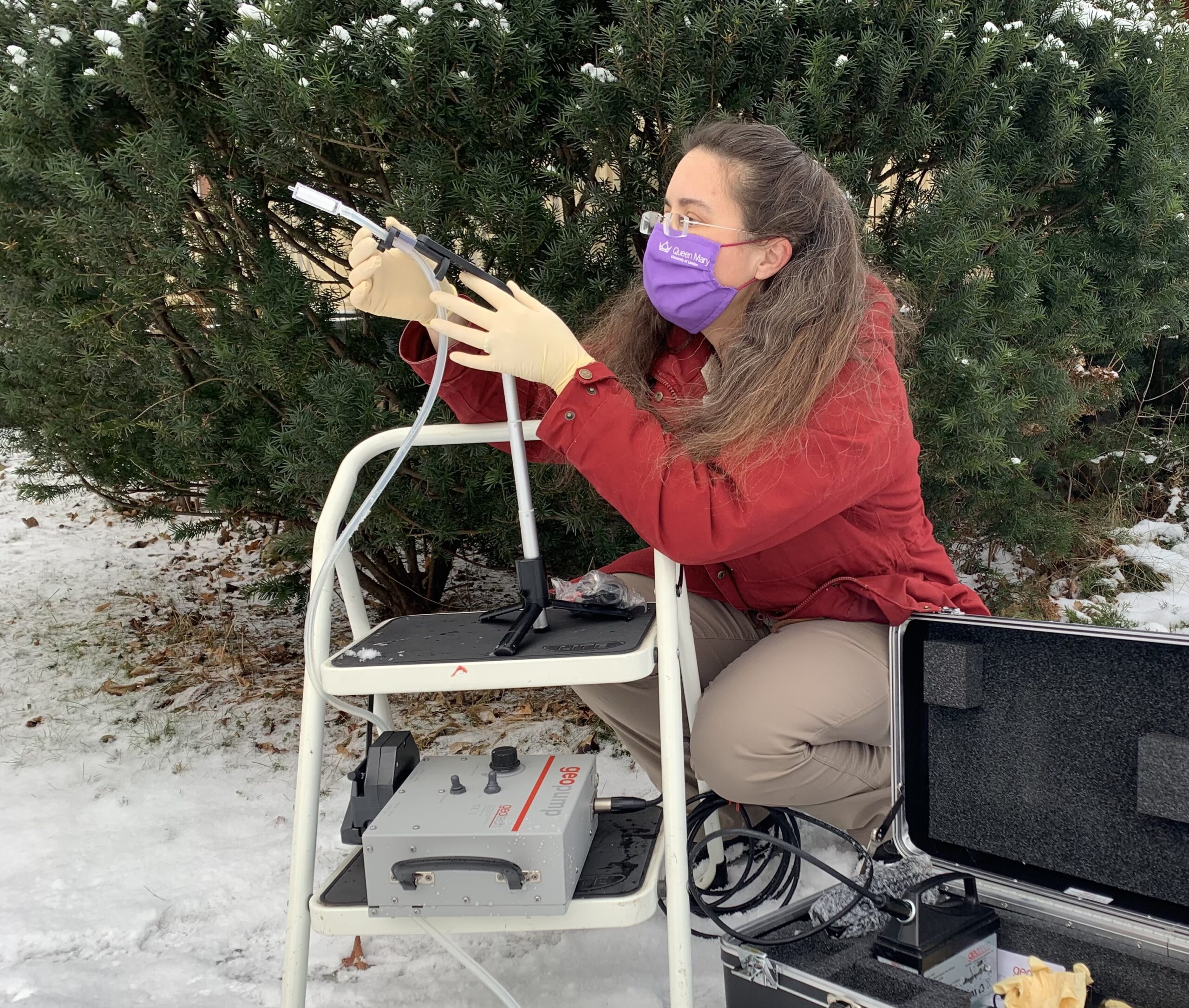 a woman wearing a red jacket and a surgical mask crouches near a bush and holds a device in the air