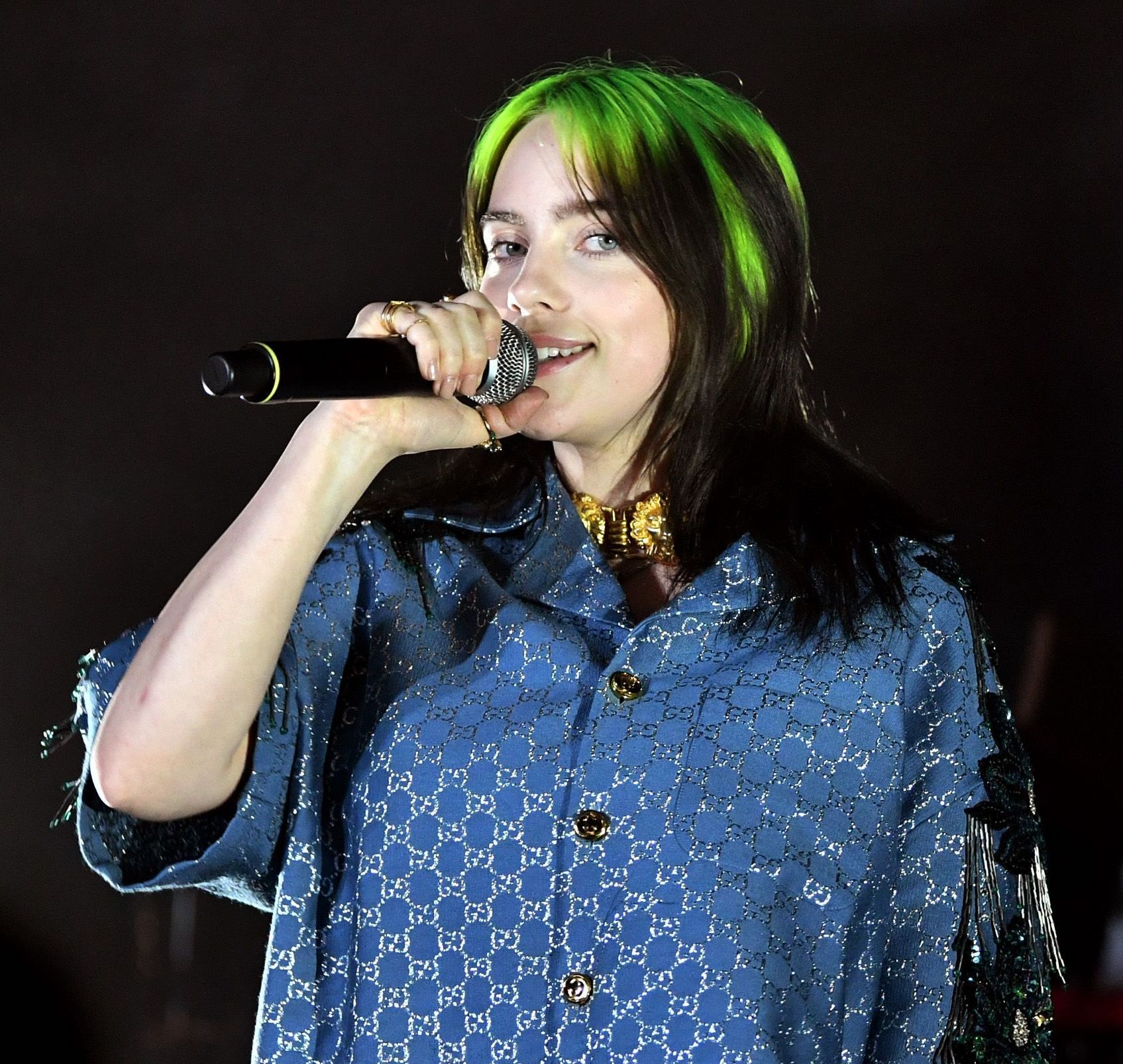 Billie Eilish, with green hair and a blue button-up shirt, stands on stage with a microphone at her mouth, smiling at the camera