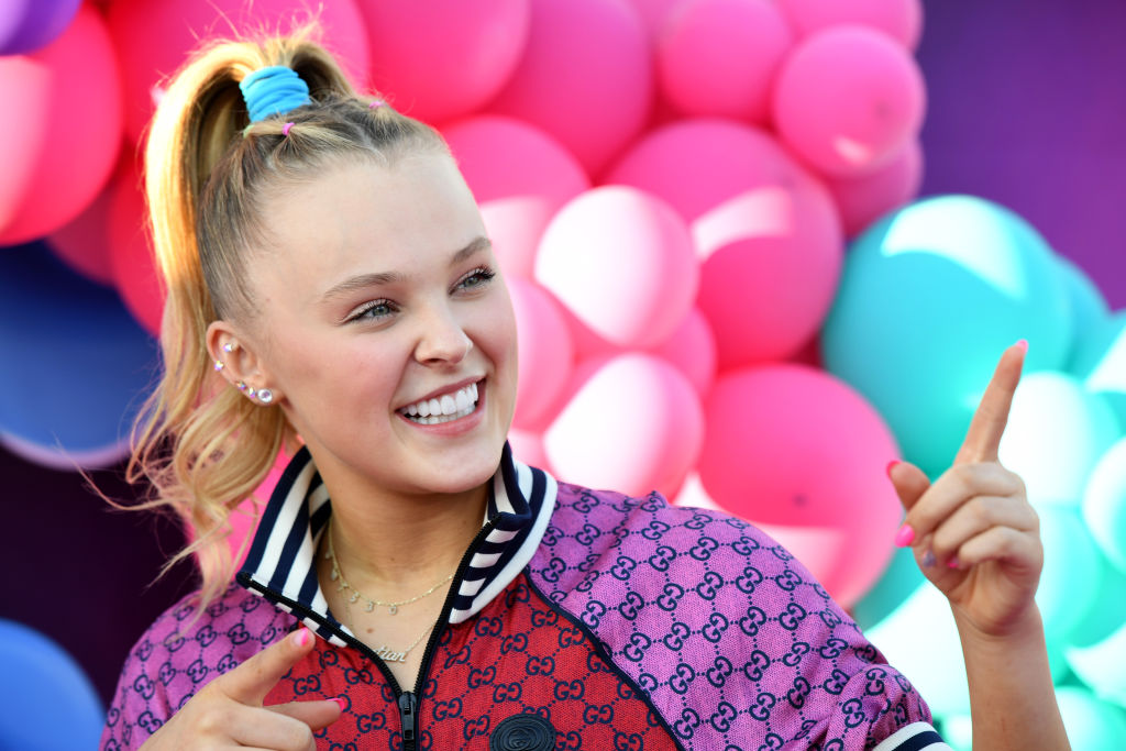 A blond girl wearing a red and purple jacket and blue hair scrunchie smiles at something off-camera