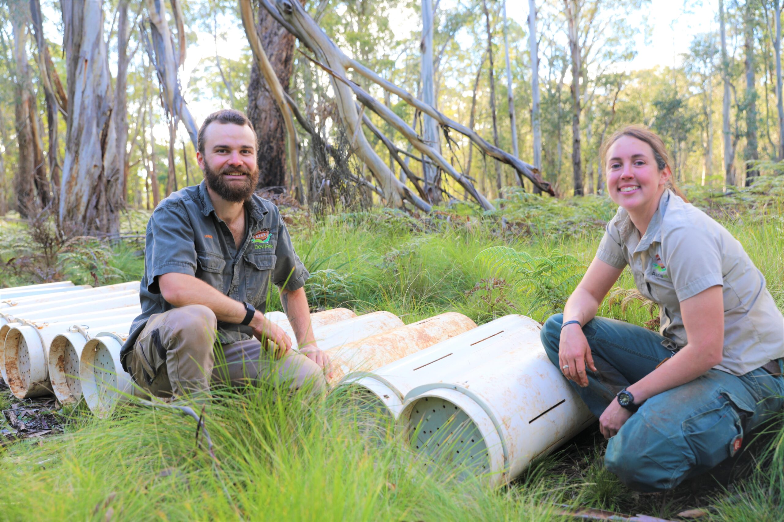 two animal keepers kneel down by cylindrical containers in a wooded area