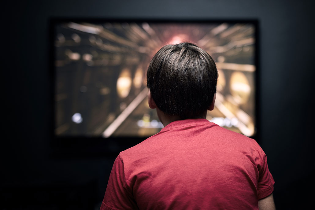 a teen with short dark hair wearing a red t-shirt is facing away from the viewer, playing a video game in a darkened room