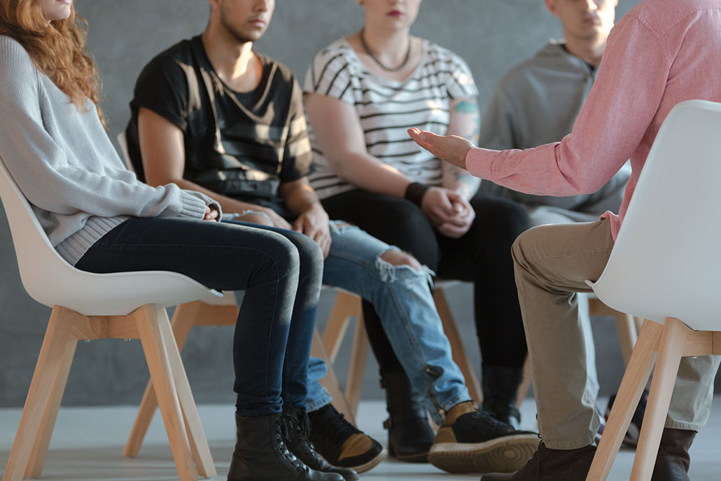 four anonymous teens sit in a group listening to a therapist speaking