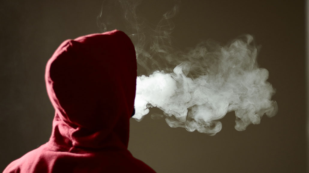 a teenager wearing a hoodie faces away from the camera and is exhaling a large cloud after vaping