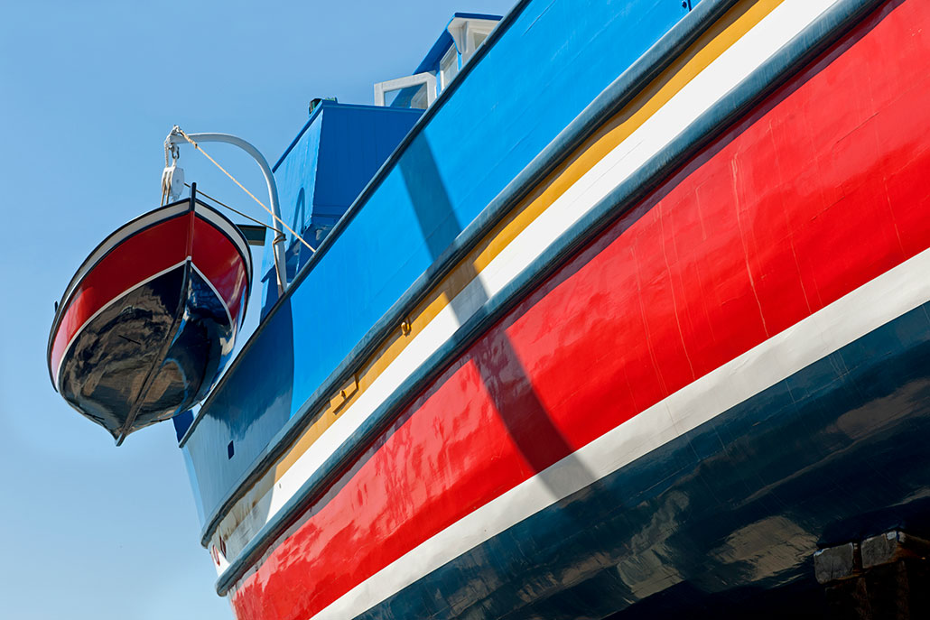 a photo of the underside of a wooden boat against a blue sky, the boat is painted in bright blue and red. a dinghy, also painted blue and red, hangs off the back of the boat