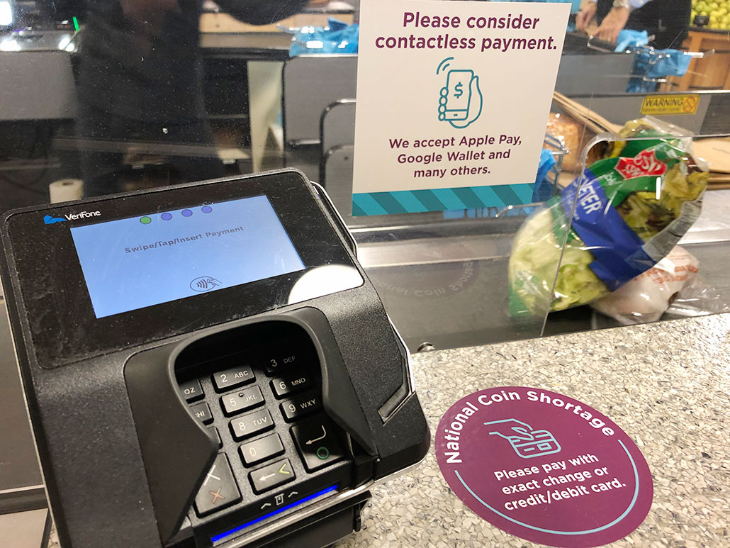a photo of a checkout line, a debit card reader machine is to the left. On the counter a circular sticker reads "National Coin Shortage: Please pay with exact change or credit/debit card." Another sticker on a clear vertical partition reads "Please consider contactless payment. We accept Apple Pay, Google Wallet, and many others."