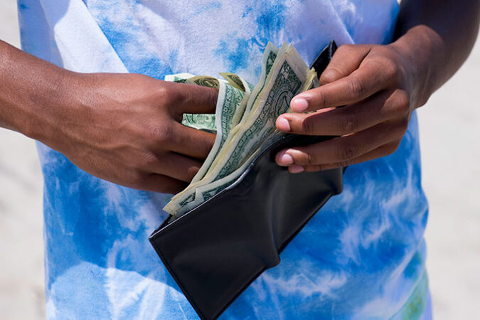 a photo of a Black teen wearing a blue and white t-shirt holding a wallet in front of their torso and removing several dollar bills from it