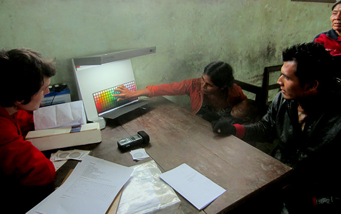 A photo of Gibson at a desk showing a Tsimane' woman a color chart. She is holding a sleeping baby in her lap and pointing to the chart. 