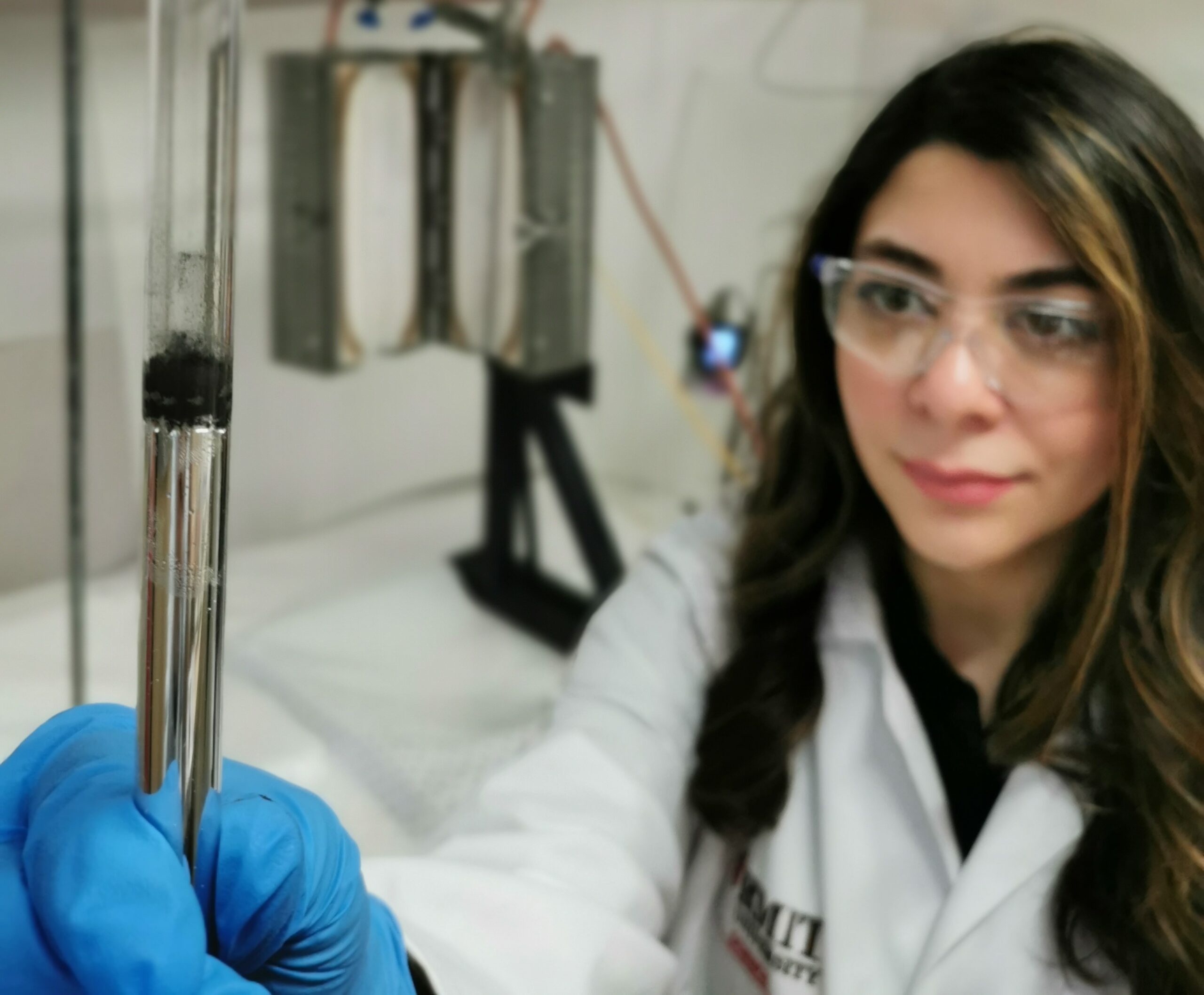 a woman wearing a lab coat and goggles holds a test tube up for the camera