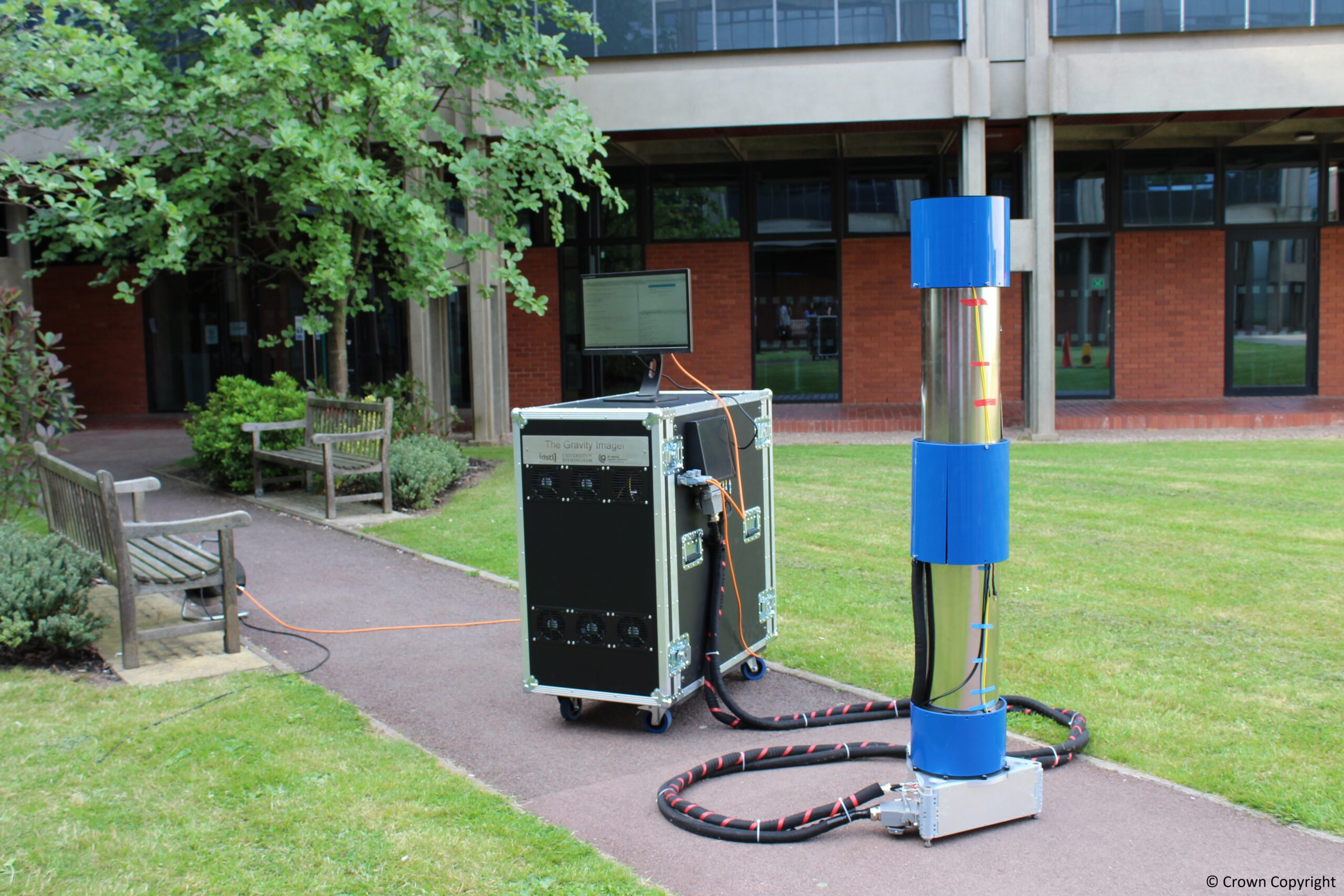 a 2-meter-tall silver cylinder capped with a blue top and bottom is tethered by cables to a cart of equipment out on a sidewalk with a building and trees in the background