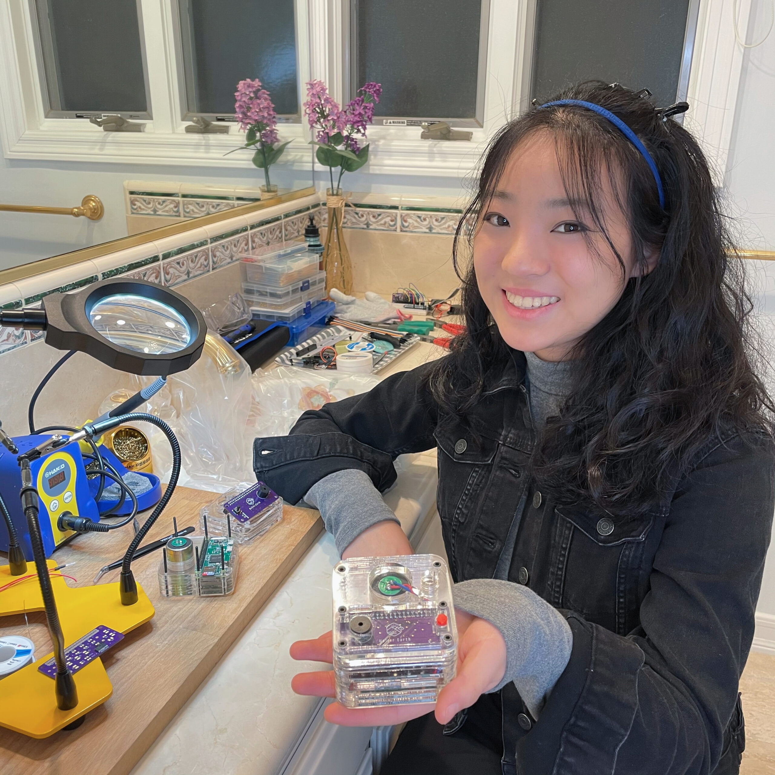 a girl sits at a bathroom counter that is cluttered with engineering materials, holding up a Rubik's cube sized device with a transparent exterior