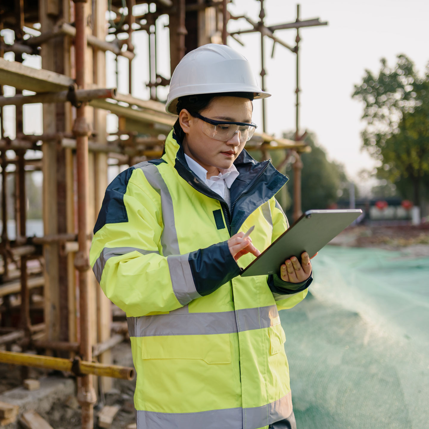 a woman wearing a hard hat and construction uniform looks at an iPad while standing in front of a partially constructed building
