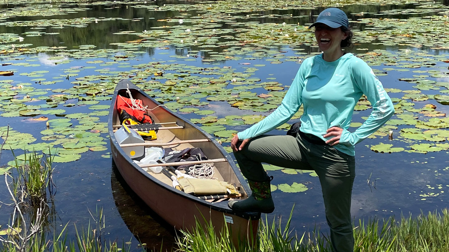 a woman wearing a ball cap, blue shirt, jeans and boots stands with one foot on a canoe parked at the edge of a pond