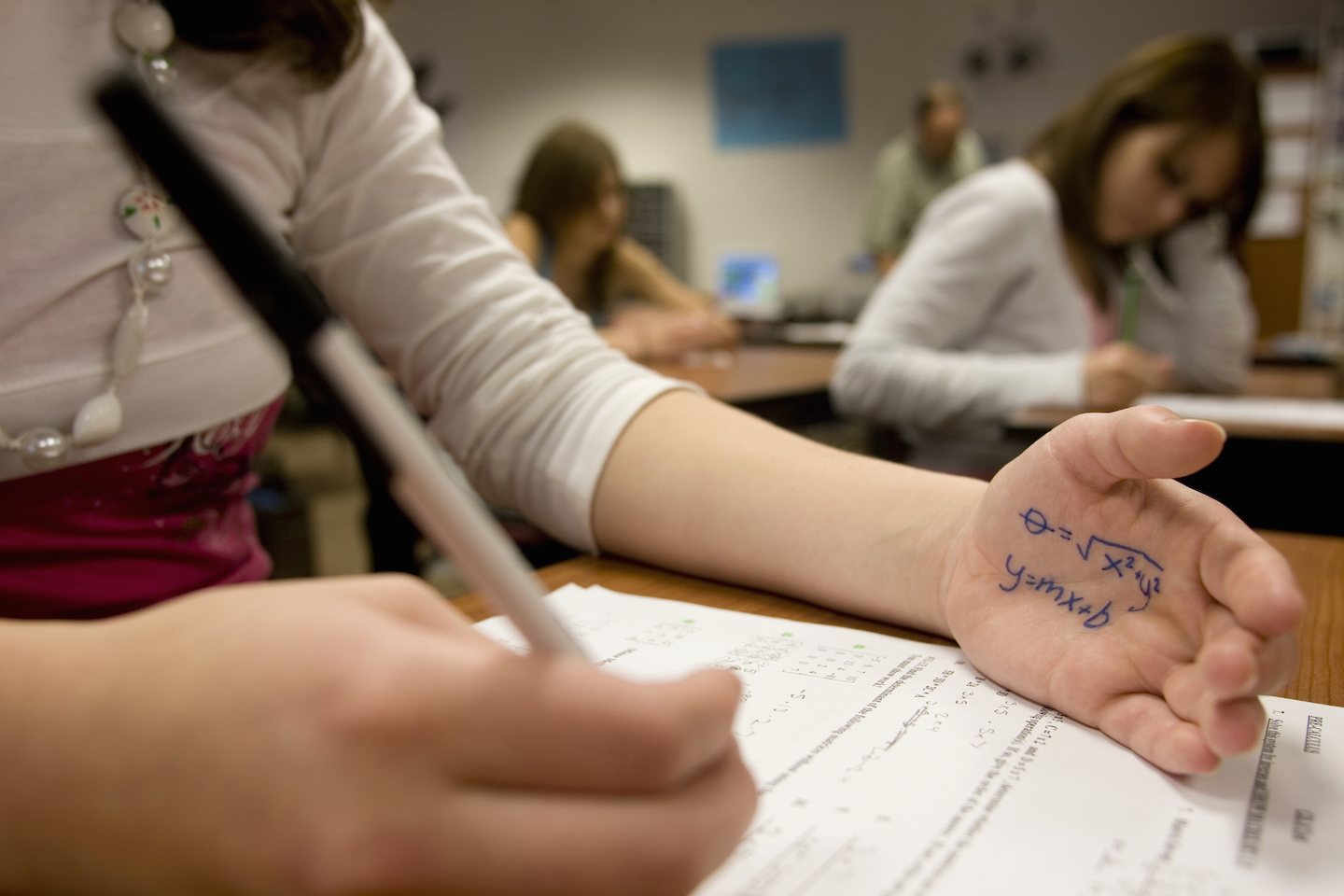 a girl taking a test holds her palm out in front of her on the desk to read an equation she has written in pen on her hand