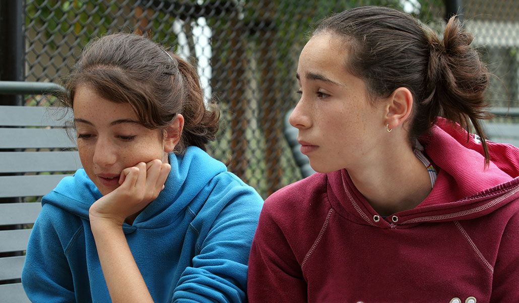 two young women with freckles, light skin, and dark hair are talking. One friend is listening with a concerned look on her face.