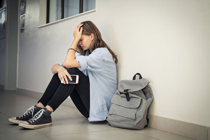 a photo of a teen sitting on a hallway floor with her back to a wall, her hand is covering her face and she looks upset