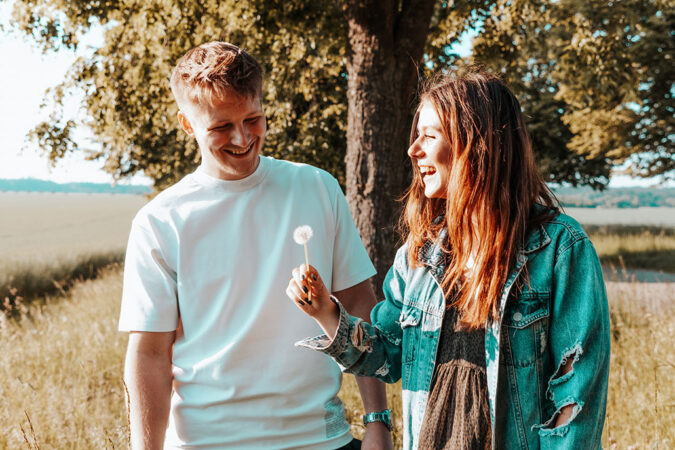 a photo of a young man and a young woman standing in a field in front of a tree. It's a sunny day and the young woman is holding a dandelion puff and laughing. The young man is smiling and looking down.
