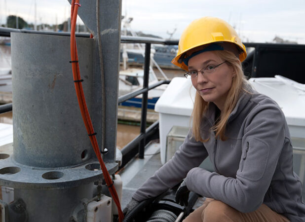 a blonde woman wearing glasses and a yellow hard hat crouches by some metal equipment outdoors