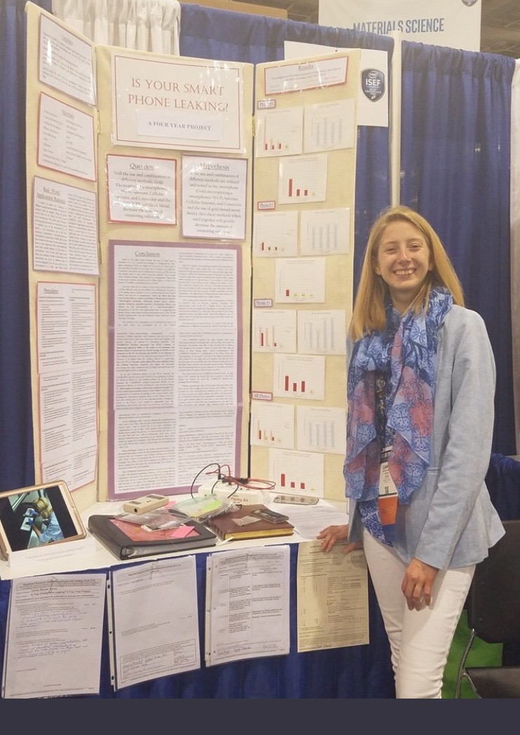 a blond teenage girl smiles while standing in front of a trifold poster