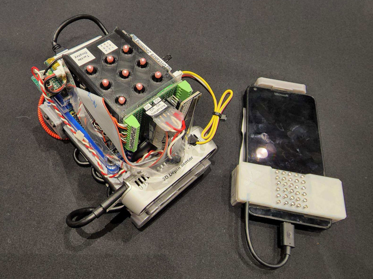 a handheld device with wires and buttons sits on a gray table beside a smartphone