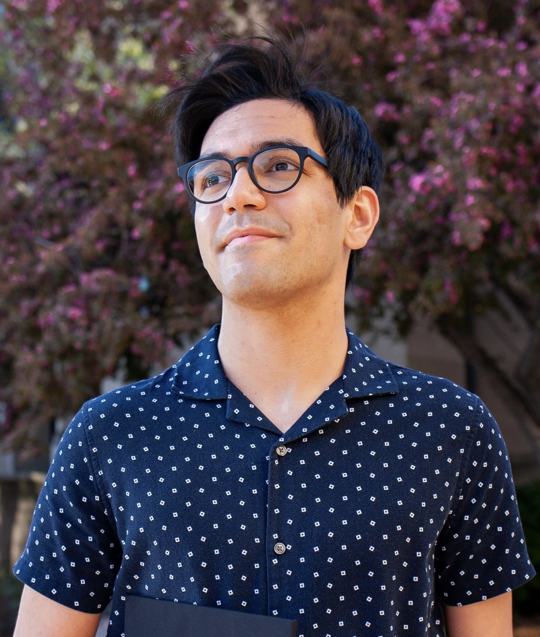 a young man wearing glasses and a blue shirt smiles while looking off-camera