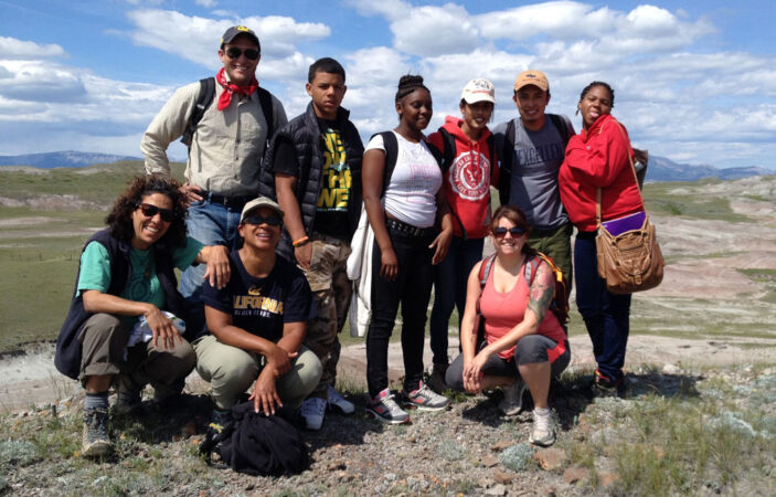 Lisa White and several college and high schol students of varying ages and backgrounds posing against a blue sky with clouds and mountains in the distance